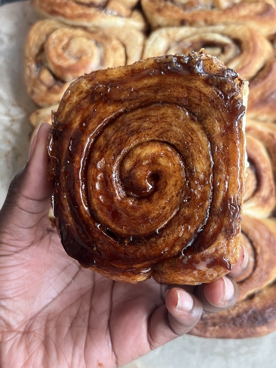 Glossy caramelised cinnamon bun held over a tray of buns